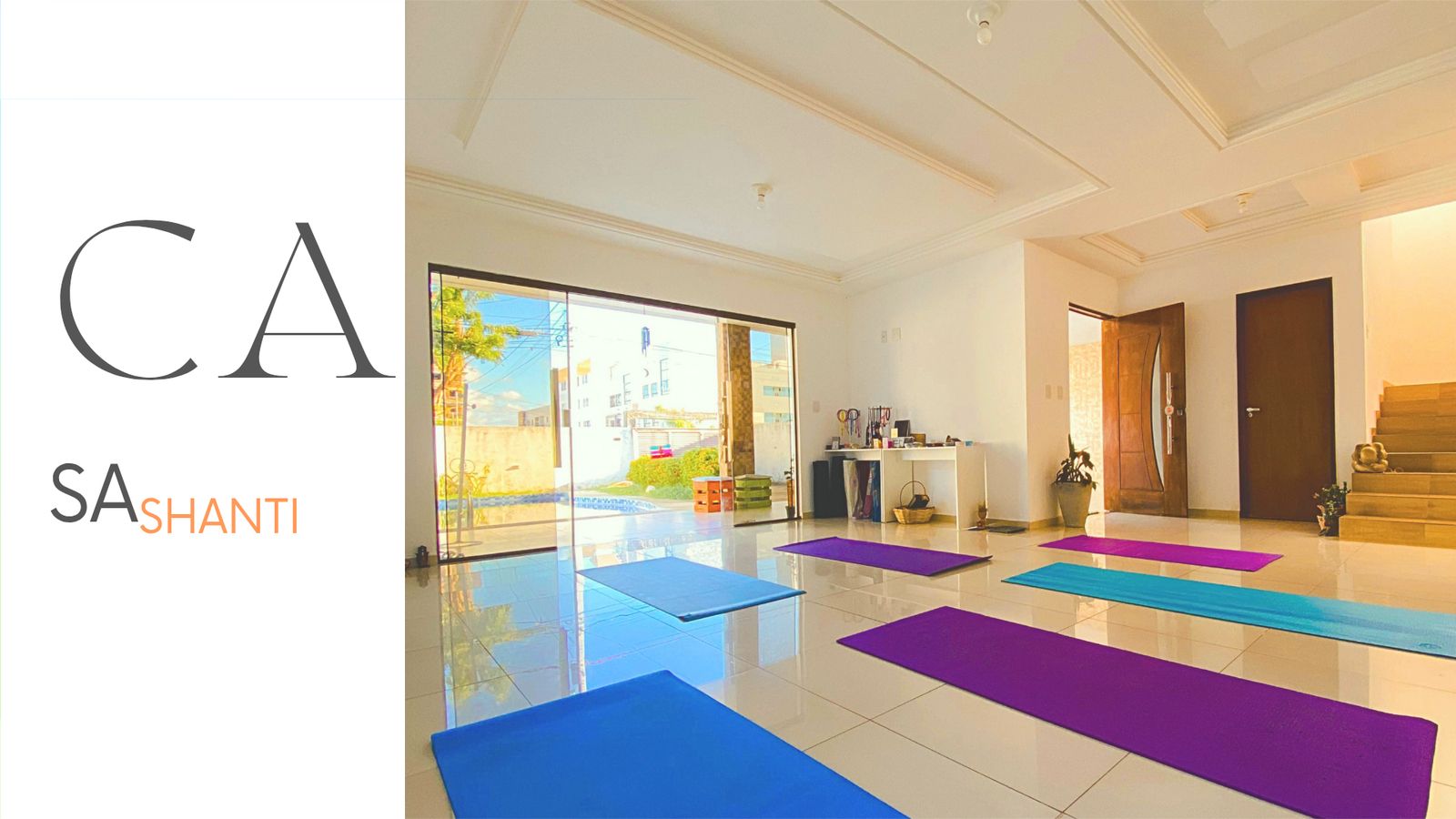 Sala ampla de um estúdio de Yoga no Bessa, em João Pessoa, com vários tapetes coloridos dispostos no chão e janelas grandes com vista para a área externa