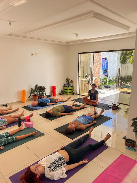 Turma de Yoga em João Pessoa praticando relaxamento (savasana) em um estúdio, com a instrutora conduzindo a aula sentada ao centro